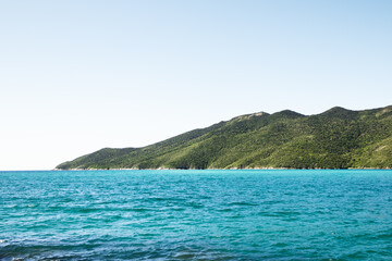 Fototapeta premium Turquoise waters meet lush green hills under a clear blue sky on a serene tropical coastline.