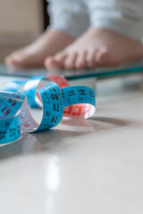 A measuring tape lying on the floor with blurred feet on a transparent scale, representing health, weight management, and fitness concepts.