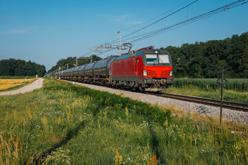 red freight train hauling cylindrical tank cars through a rural landscape. It s a sunny day with clear skies, green fields, wildflowers, and a nearby dirt path beside the tracks.