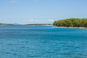 breathtaking view from Draga Bay, gazing towards Pakostane, where crystal-clear waters meet a distant shoreline framed by pine trees. The serene seascape shines under a vast, partly cloudy sky, a