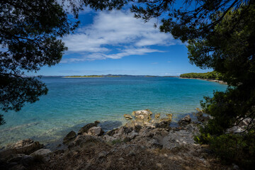 breathtaking view from Draga Bay, gazing towards Pakostane, where crystal-clear waters meet a distant shoreline framed by pine trees. The serene seascape shines under a vast, partly cloudy sky, a