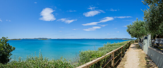 scenic path winds alongside a wooden fence towards Pakostane, overlooking the stunning turquoise waters and distant islands under a bright, partly cloudy sky. A perfect stroll in nature's embrace.