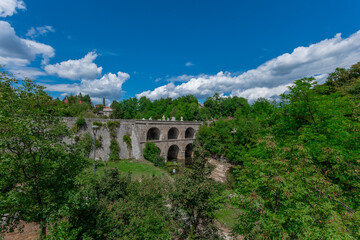The majestic Jozefinski Most na Tounjcici arches gracefully over a lush valley, its historic stone structure framed by vibrant greenery under a brilliant blue sky with scattered clouds, a timeless