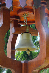 A light bronze bell set into a dark, ornate wooden frame. The background shows blurred green foliage