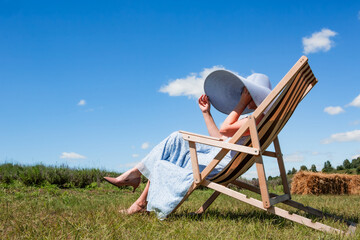 Young beautiful sexy woman in a big hat sits on a sun lounger on a meadow on a sunny day against the sky. Rest. Vacation. Tourism.	