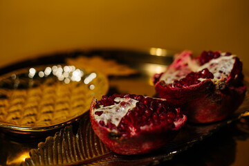 Two juicy halves of a pomegranate are placed on a golden tray on a colored background near a green branch of the plant. A composition for interior decoration and a festive table.