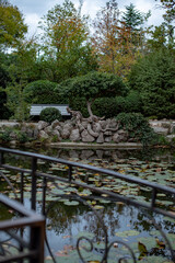 A tranquil park scene featuring a pond with lily pads, surrounded by lush greenery and a stone...