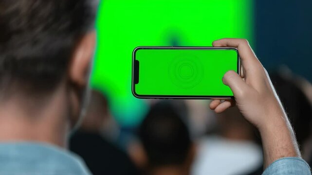 Person holding smartphone with green screen in hand, capturing video or vdo at event, surrounded by blurred audience, technology and communication concept, modern lifestyle, focus on device