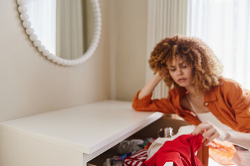 Frustrated woman with curly hair searching through messy drawer in a modern, bright room, showcasing emotions of confusion and stress over clothing choices