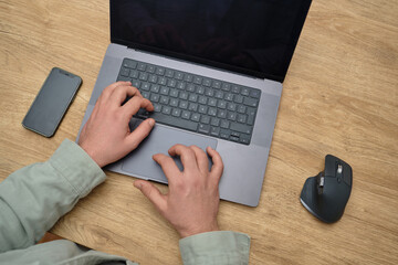 Close-up of a person typing on a laptop keyboard at a wooden desk, with a smartphone and ergonomic mouse nearby. Productive workspace setup.