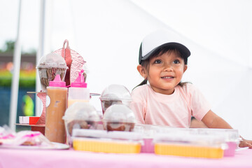 Smiling three-year-old Colombian girl seated behind pink table full of pastries at entrepreneur fair
