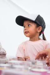 Portrait of three-year-old Colombian girl wearing cap at kids’ entrepreneurship fair, bright setup