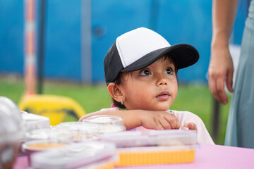 Curious three-year-old Colombian girl sampling treats at kids’ entrepreneurship fair, front view