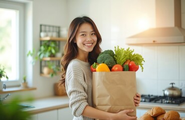 Young Asian woman stands in cozy kitchen holding paper bag filled with fresh produce. Brown sweater, warm smile, white tile backsplash, stove, counter with kitchen appliances. Fresh food, groceries,