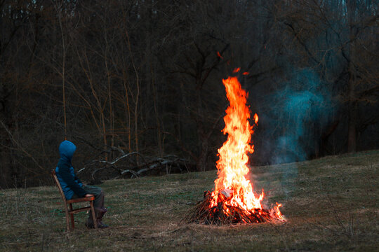 A man in a hooded jacket sits by a campfire outdoors in the autumn evening. View from the side. Travel and outdoor recreation concept.