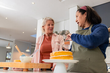 Diverse senior female friends spreading frosting together on cake at modern kitchen countertop