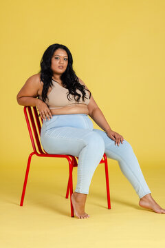 Indian woman sitting on red metal chair against yellow backdrop in studio wearing jeans barefoot