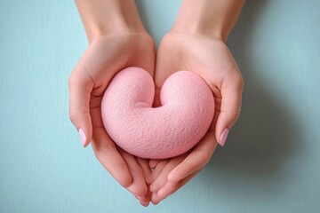Obraz premium Two hands gently holding a soft pink heart-shaped object against a light blue background, evoking care and tenderness