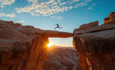 Fototapete Person leaping across a natural rock arch during sunset with dramatic sky and expansive rocky landscape © dgete