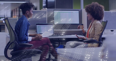 Discussing two women wearing smart clothes, gesturing with coffee cup, typing on laptop in office