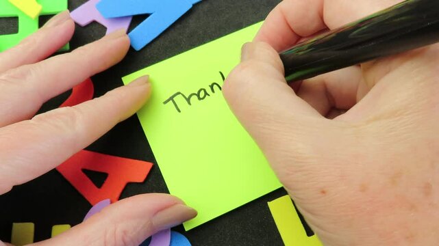 Woman hands writing the expression "Thank you Teacher" with a smile on a yellow greeting card among multicolored eva foam letters. Black background. Teacher's Day celebration concept.