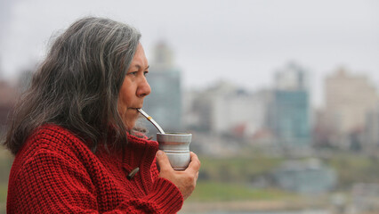 Close-up portrait of an elderly Latin American  woman with Guaraní features, wearing warm knitted clothes and drinking  a mate outdoors. She looks calm and natural in a peaceful setting.