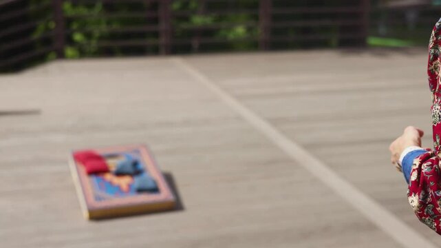 Cornhole game set, process of throwing bean bags, female girl tossing bean sacks, corn hole in the backyard, wooden boards for corn-hole tournament in a summer day on a wooden flooring terrace deck