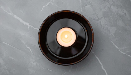 Obraz premium Overhead view of a lit candle in a dark ceramic bowl on a marble surface.