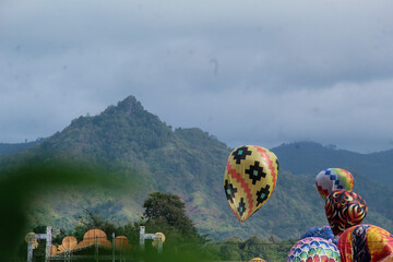 Fototapeta premium Traditional hot air balloons rise against a scenic mountain backdrop during a cultural festival in a rural area.
