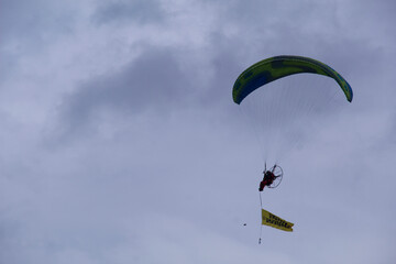 A powered paraglider flying in the sky while carrying a promotional banner during a public event.