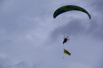 A powered paraglider flying in the sky while carrying a promotional banner during a public event.