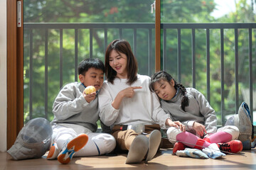 The fencers' families rest on the floor as they wait for the next round of the competition. Mother, son and daughter sit against a glass wall under the warm sunlight.