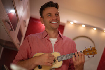 Smiling man in a pink shirt playing ukulele in a cozy modern kitchen, enjoying music, relaxing, and...