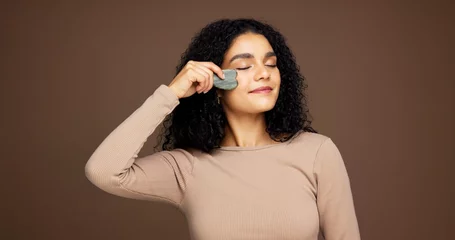 Fotobehang Beauty, skincare and woman with gua sha in studio for facial care, detox and lymphatic drainage. Dermatology, spa and person with jade tools for anti aging, massage or smooth skin on brown background © peopleimages.com