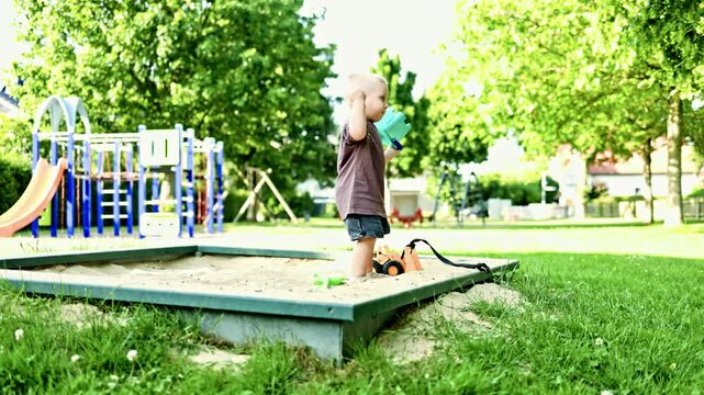 Cute blond boy 2 years old in a t-shirt and shorts on a summer walk on the playground. Toddler digs sand with a shovel in the sandbox. Summer games on the playground outdoors.