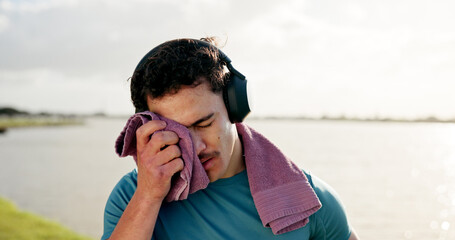 Tired man, sweat or fitness with towel or headphones at lake for intense workout, break or recovery. Active, male person or dry with cloth for physical activity, exercise or outdoor training at river