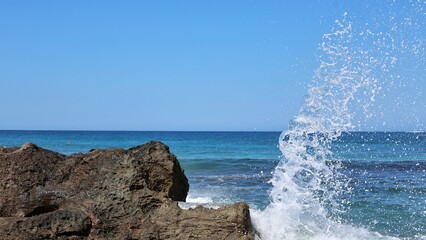 Panoramic view of a sea wave crashing against volcanic rocks on the Crete coast under a clear blue...