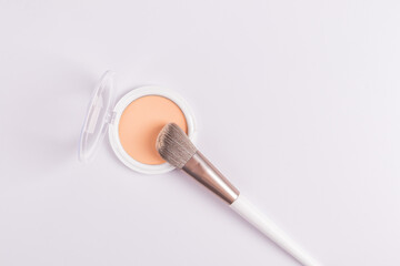 Open white round pack of compact powder in natural tone on a white background with a makeup brush....
