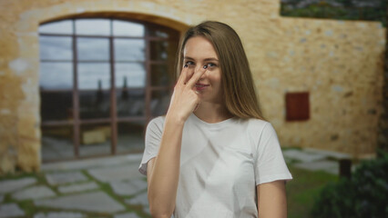 Young woman outdoors gesturing with a finger near her eye, posing against a stone wall with closed eyes and a relaxed smile, conveying a sense of observation and mindfulness.
