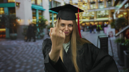 Graduated young woman in cap and gown on a busy city street shows a playful expression amidst urban...
