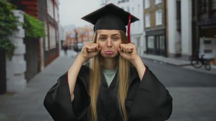 Young woman in graduation gown mockingly gestures crying on a city street, blending humor and...