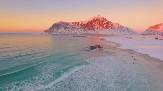 A stunning aerial view of a snowy beach with gentle waves and majestic mountains under a beautiful pink sunset sky during winter. This high-quality footage captures the serene and tranquil beauty of a