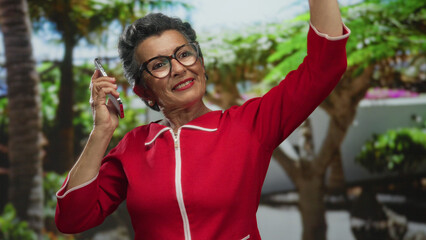 Fototapeta premium Senior woman with grey hair, wearing red dress, dances joyfully listening to music on smartphone in a vibrant green park, surrounded by lush trees and plants.