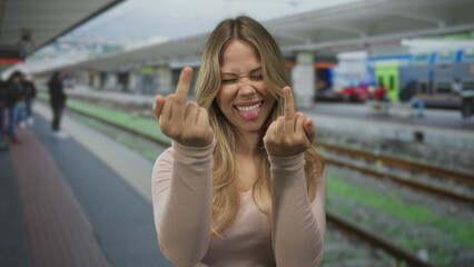 Young caucasian woman flips both middle fingers and sticks tongue out at crowded train station...