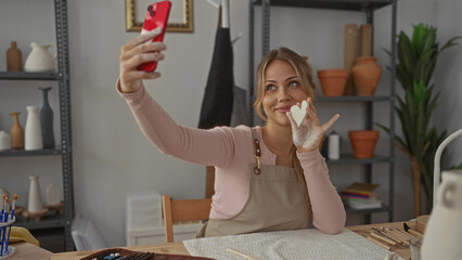 Woman holds clay heart while raising smartphone for selfie in a pottery studio; creativity selfexpression joy.