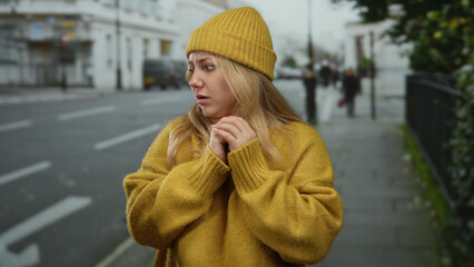 Woman in yellow sweater and hat looks anxious on city street, capturing emotion of fear in urban...
