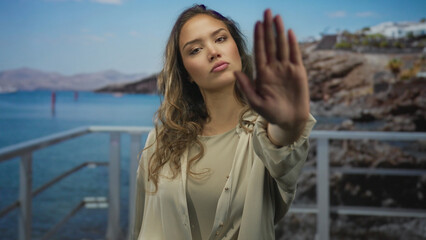 Woman with serious expression extends palm for stop on seaside promenade; defiance refusal boundary.