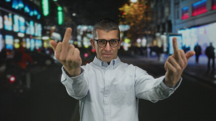 Man with glasses making offensive gesture on illuminated city street at night showing defiance and...