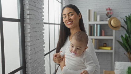 Woman smiling holding baby in cozy living room with white brick wall and bookshelves, sunlight streaming through big windows, portraying warmth, love, and family bonding indoors.