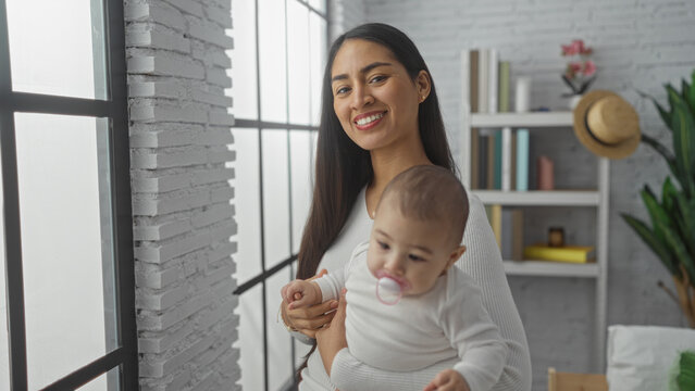 Woman smiling holding baby in cozy living room with white brick wall and bookshelves, sunlight streaming through big windows, portraying warmth, love, and family bonding indoors.